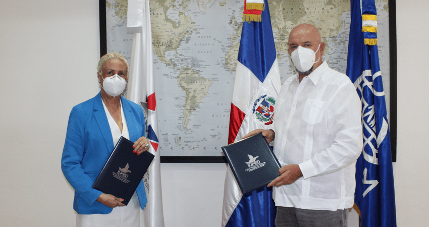 Representantes de OIM e INM firman convenio. Posan frente a mapamundi y banderas
