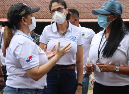 Mujeres con mascarilla conversan. Sus camisetas tienen logos de la OIM y el gobierno de Panamá. 