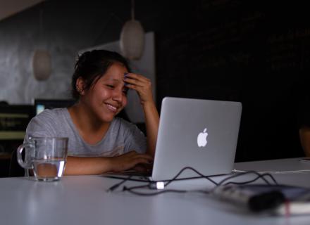 Mujer morena joven sonríe frente a pantalla de computadora. Está en una casa y tiene un vaso de agua. 